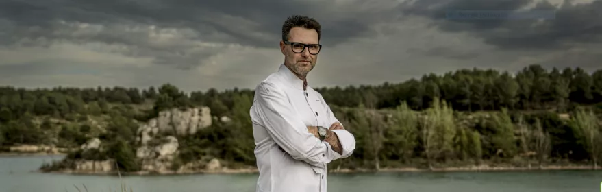 Chef con chaquetilla blanca posando junto a un paisaje natural y un río.