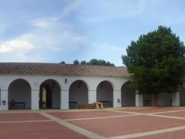 Vista panorámica de una plaza porticada con edificios blancos y árboles.