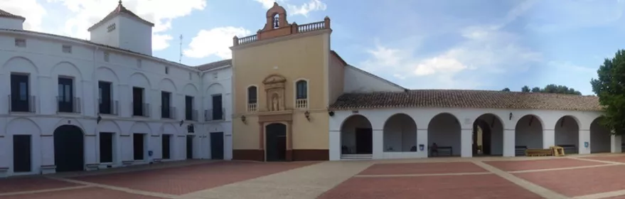 Vista panorámica de una plaza porticada con edificios blancos y árboles.