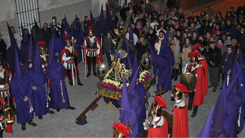 Procesión nocturna con paso religioso rodeado de penitentes y escolta ceremonial, ante numeroso público.