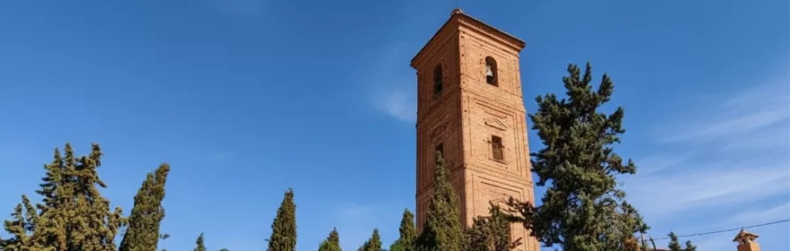 Torre de ladrillo rodeada de pinos y edificios encalados en día soleado.