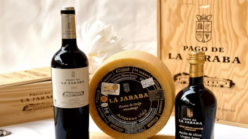 Botellas de vino, queso curado y aceite sobre una tabla, con cajas de madera de fondo.