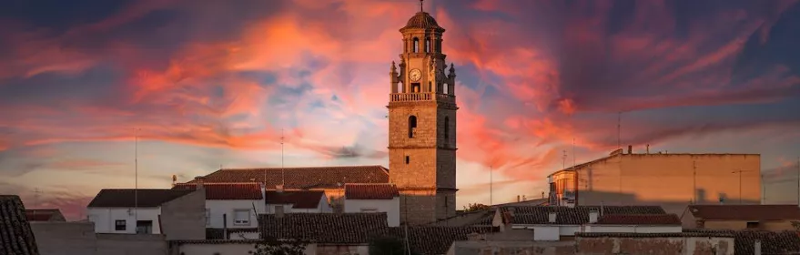 Torre de la iglesia de La Gineta destacando sobre los tejados del municipio durante un atardecer de tonos rosados.