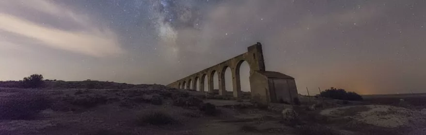 Antiguo acueducto de La Herrera bajo un cielo nocturno estrellado con la Vía Láctea visible sobre el paisaje manchego.