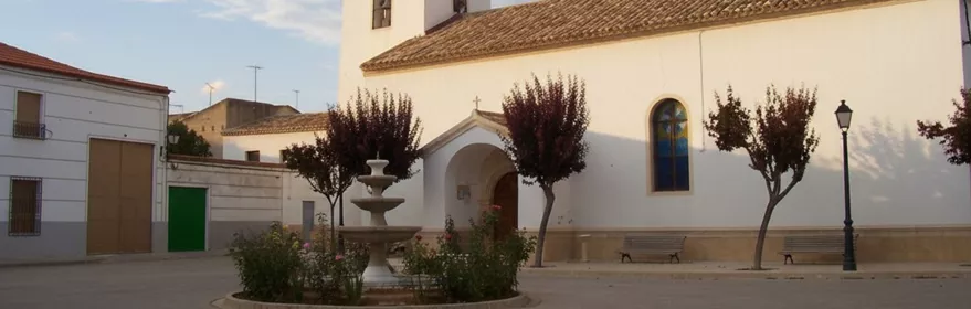 Iglesia de Montalvos con fachada blanca y torre cuadrada en una plaza tranquila con fuente central.