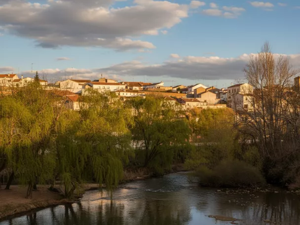 Vista de Villalgordo del Júcar junto al río, con árboles y casas iluminadas al atardecer.
