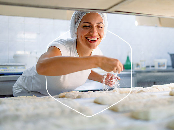 A joyful female baker in white attire and hairnet sprinkles flour on dough pieces, showing excitement while preparing them for baking in a professional kitchen setting.
