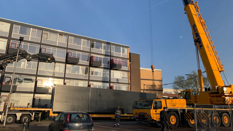 Crane getting ready to lift a prefabricated elevator from a truck in front of a residential building.