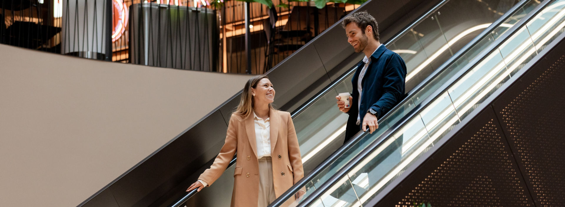 A young man and woman in professional clothes talk on escalator in a shopping center.