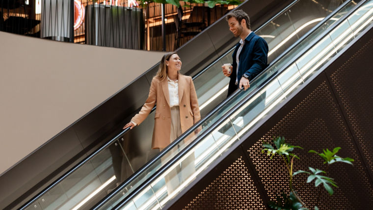 A young man and woman in professional clothes talk on escalator in a shopping center.
