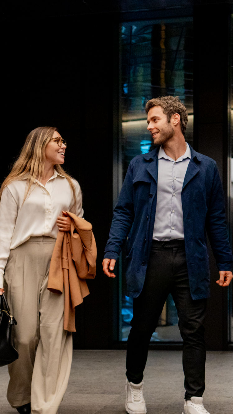 A man and a woman in casual business wear walk towards an elevator while talking happily.