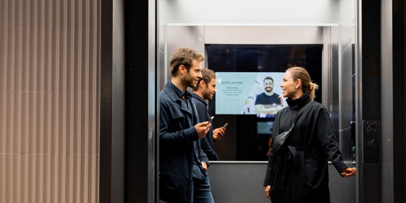 KONE - Two people in an elevator conversing with each other, while other is pressing floor button.