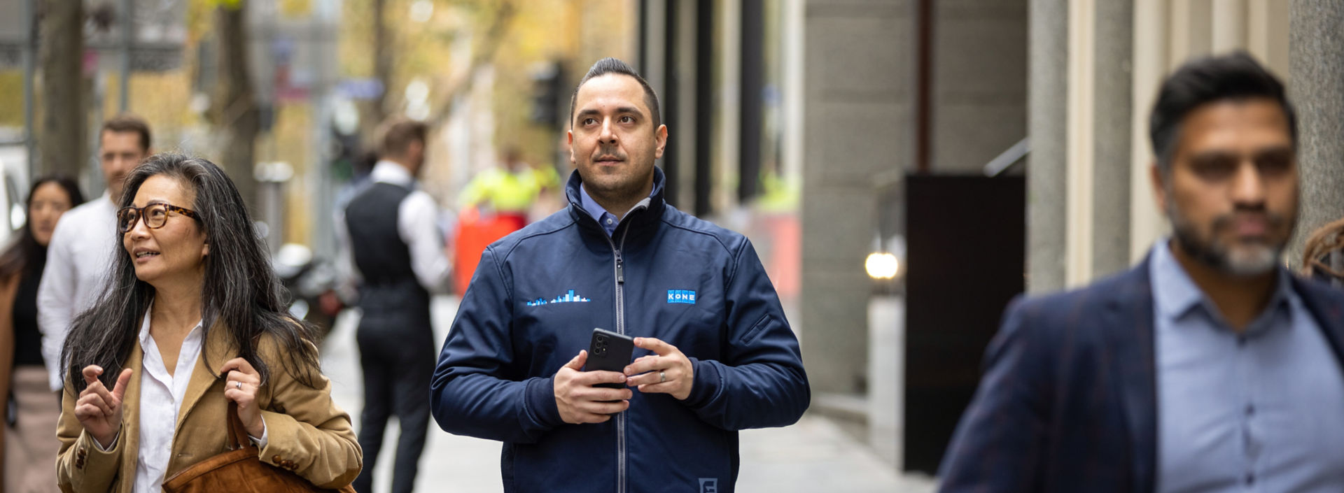 A KONE technician walks down a busy urban street holding a mobile device.