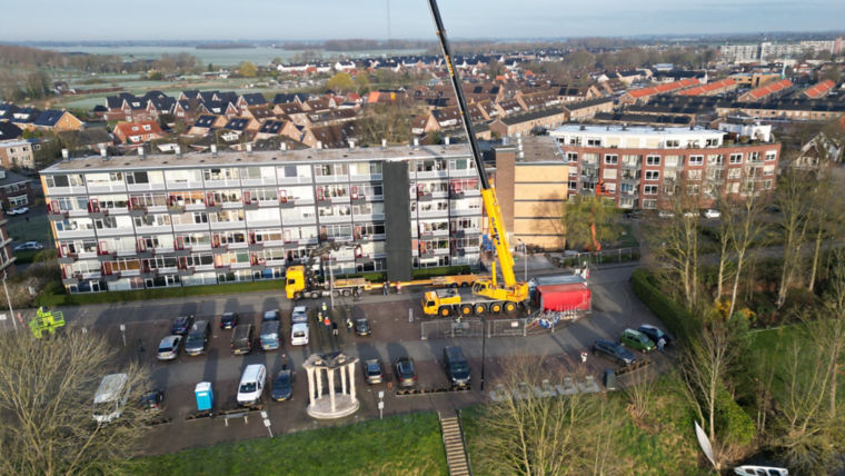 Crane getting ready to lift a prefabricated elevator from a truck in front of a residential building.