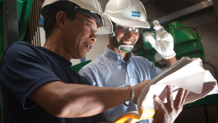 Two KONE colleagues in hard hats smile while looking at a document.