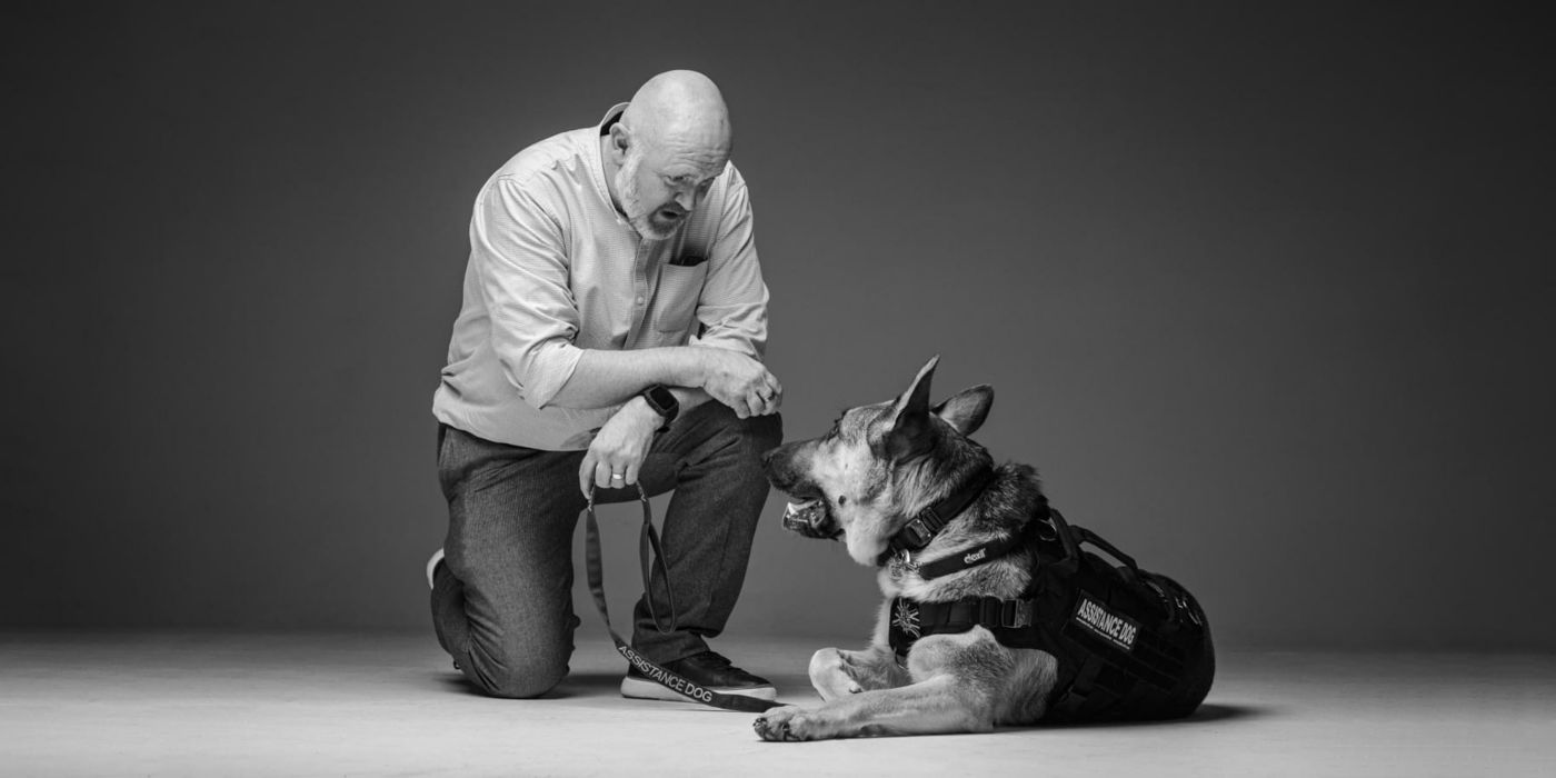 Man kneeling and looking at his German Shepherd wearing an assistance dog vest.