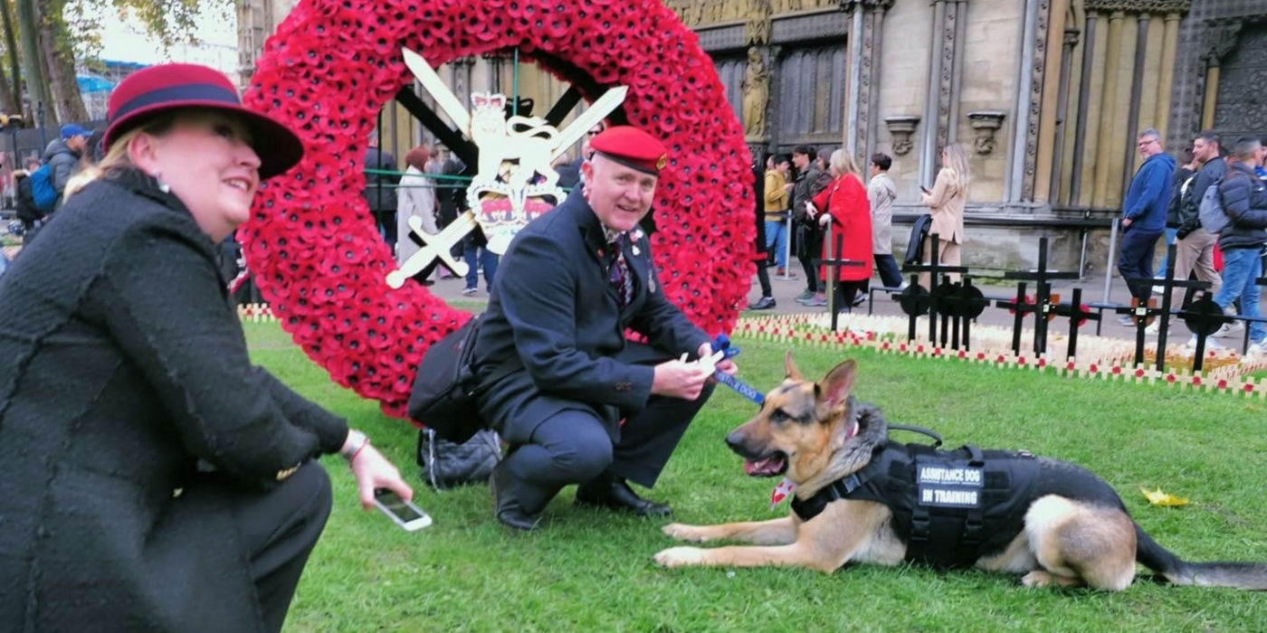 Man and woman crouching on grass with their German Shepherd next to a red poppy wreath.
