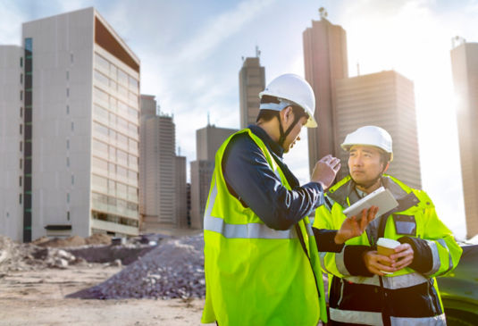 Two men in hi-vis jackets have a conversation on a building site while looking at a tablet.