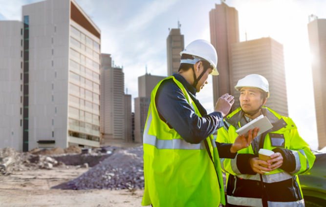Two men in hi-vis jackets have a conversation on a building site while looking at a tablet.