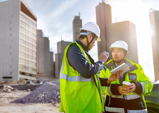 Two men in hi-vis jackets have a conversation on a building site while looking at a tablet.
