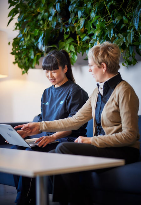 Two KONE employees working in front of a computer while sitting on a couch and conversing with each other.
