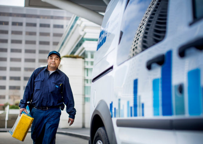 Técnico KONE con su maletín de trabajo caminado afuera de un edificio en dirección a su vehículo KONE