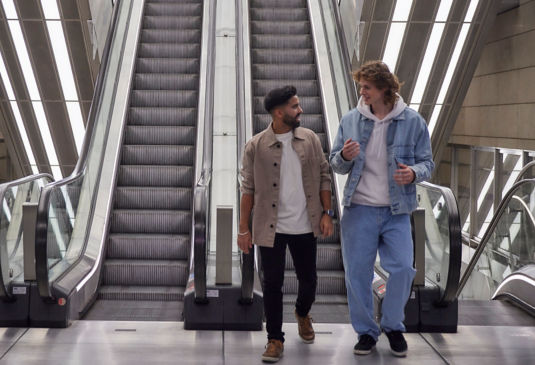 Two men walking from an escalator while having a conversation.