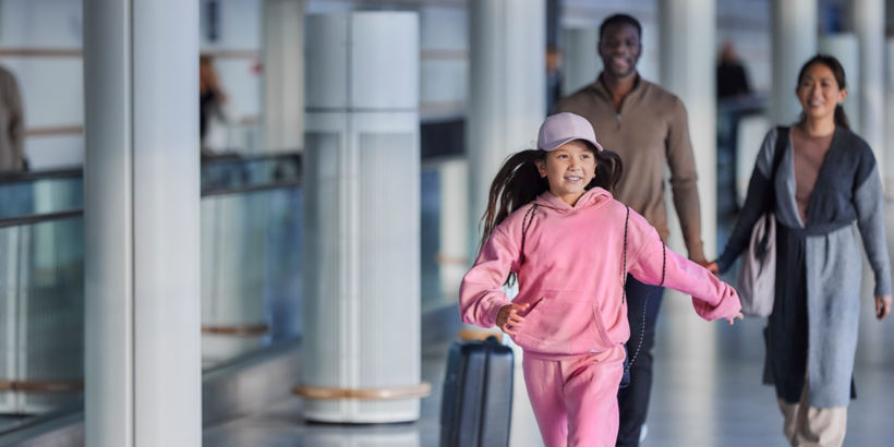 En glad jente som løper på flyplassen med foreldrene sine og en rulletrapp i bakgrunnen – KONE rullebånd, KONE rulletrapp.
