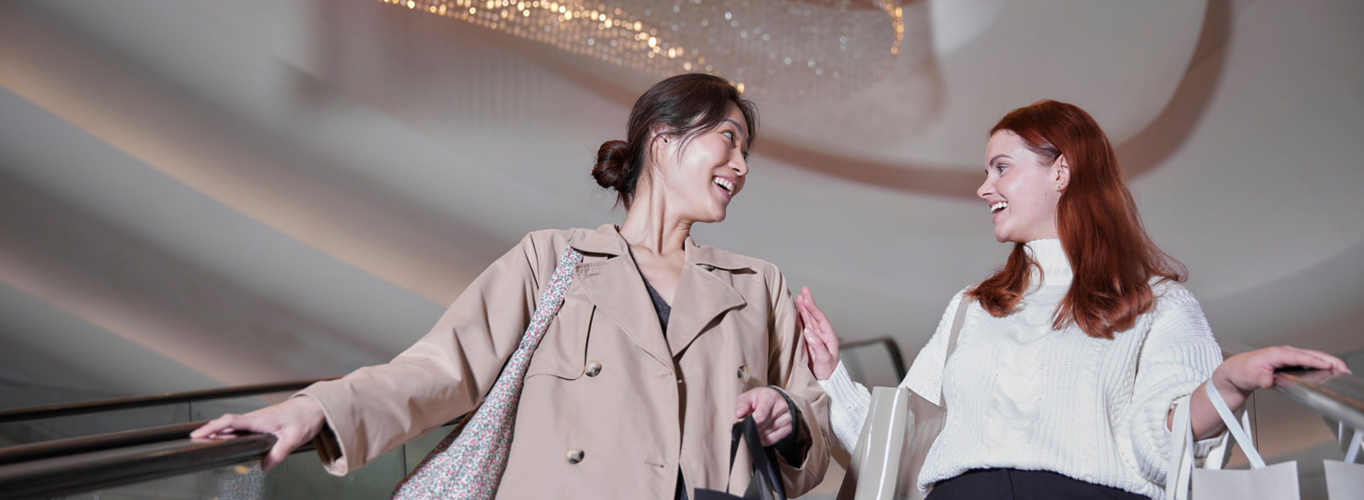Two women on an escalator with shopping bags at a modern mall