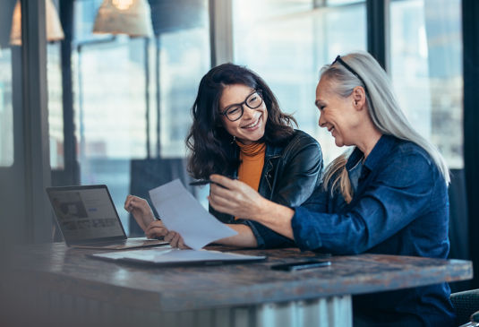 Two women conversing over a table while holding paper document in hand.