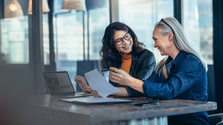 Two women conversing over a table while holding paper document in hand.