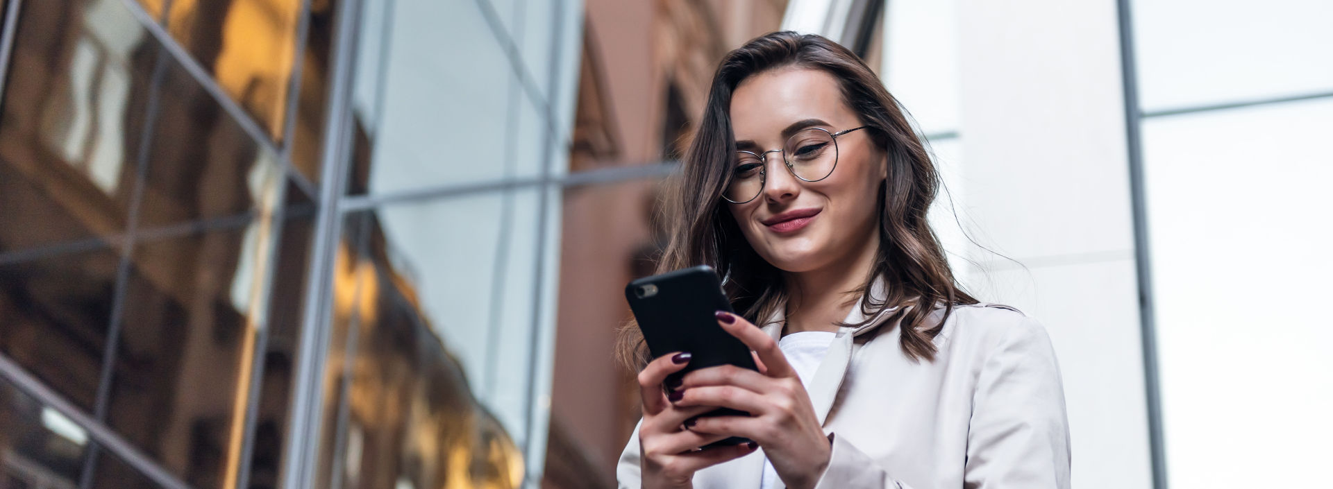 A smiling, curly-haired young woman wearing trendy sunglasses walks down a city-centre street and uses her phone.