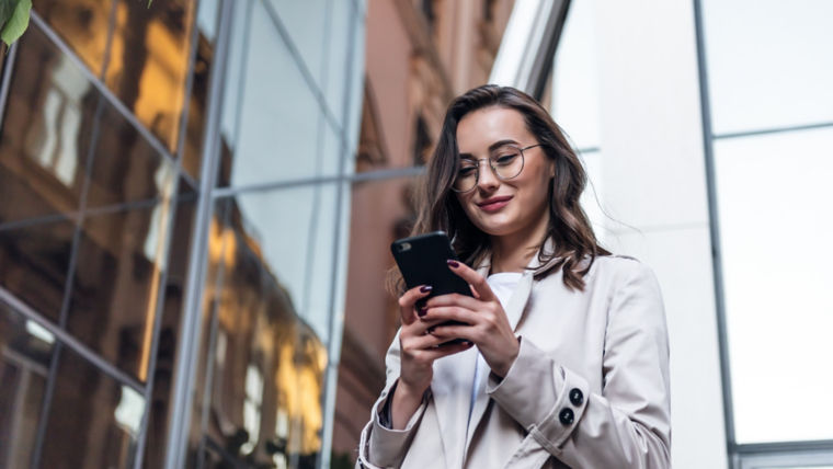 A smiling, curly-haired young woman wearing trendy sunglasses walks down a city-centre street and uses her phone.