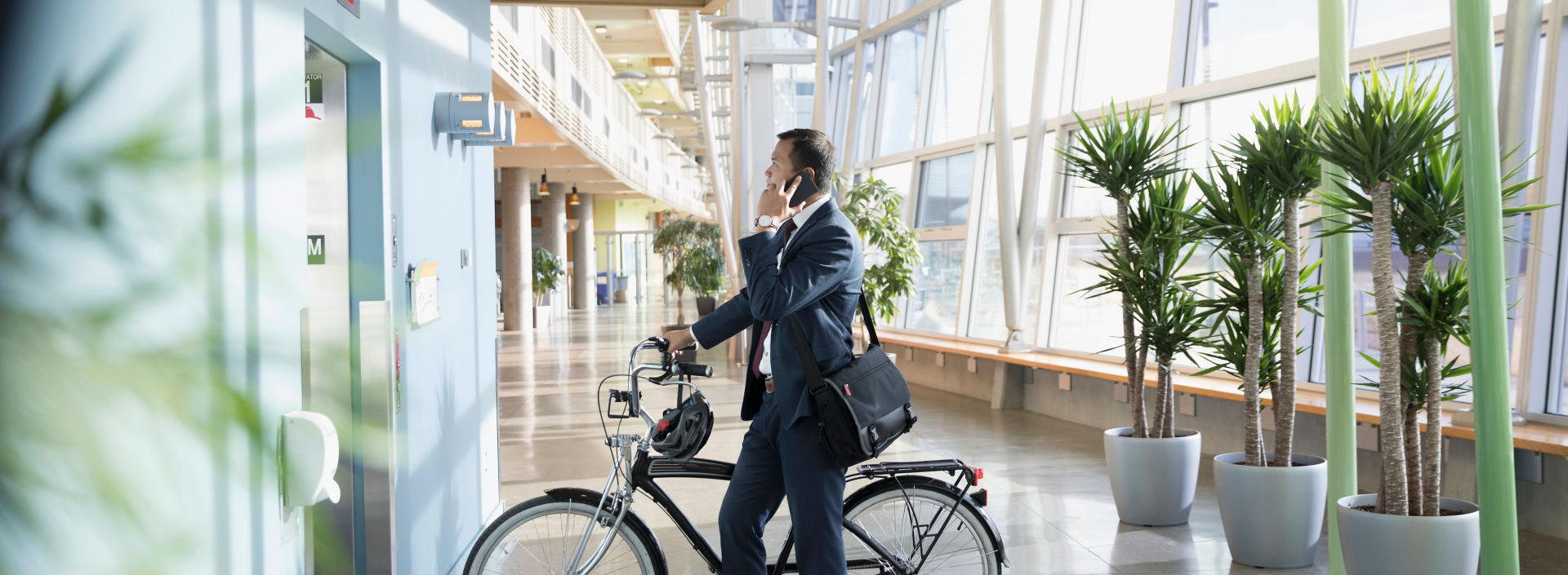 Businessman commuter with bicycle talking on smart phone, waiting at elevator in office lobby.