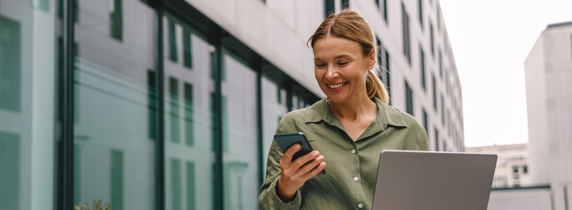 Woman sitting outside next to office building, working with computer and checking smartphone.