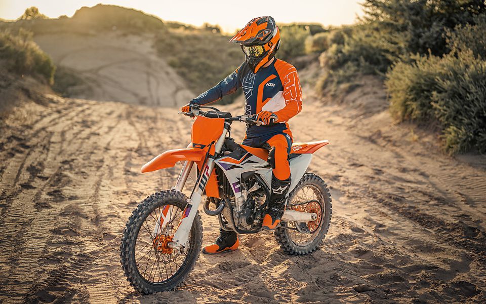 A men with a KTM Backpack on his back, standing next to a Rider wearing orange Racing clothes, sitting on a KTM Motocross Bike.