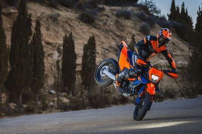 Motorcyclist in orange gear performs a wheelie on a KTM Enduro Bike over a rock in a hilly landscape under clear blue sky.