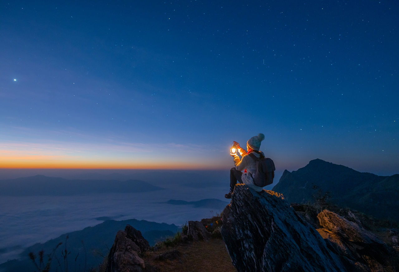 Person on a mountain ridge at twilight with a light, overlooking a mountainous landscape.