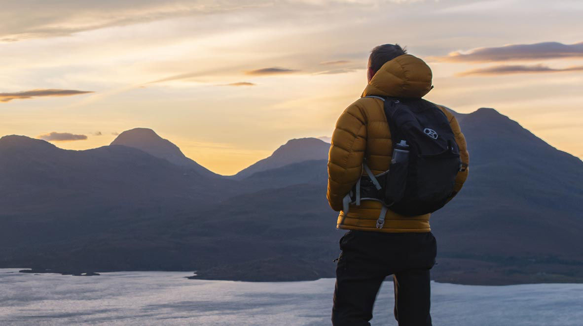 Hiker standing above a lake with mountains in the distance under an evening sky.