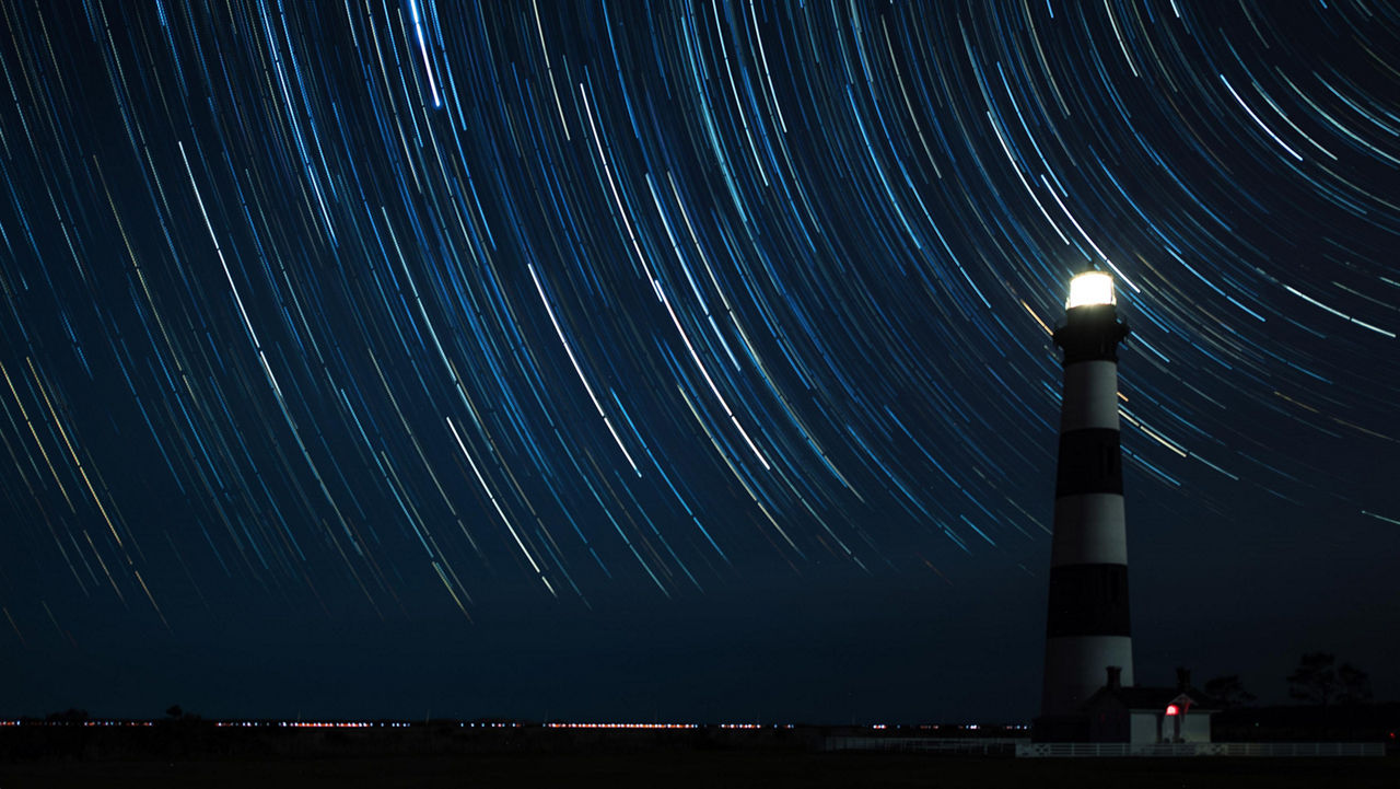 Lighthouse illuminated at night beneath circular star trails in a dark blue sky.