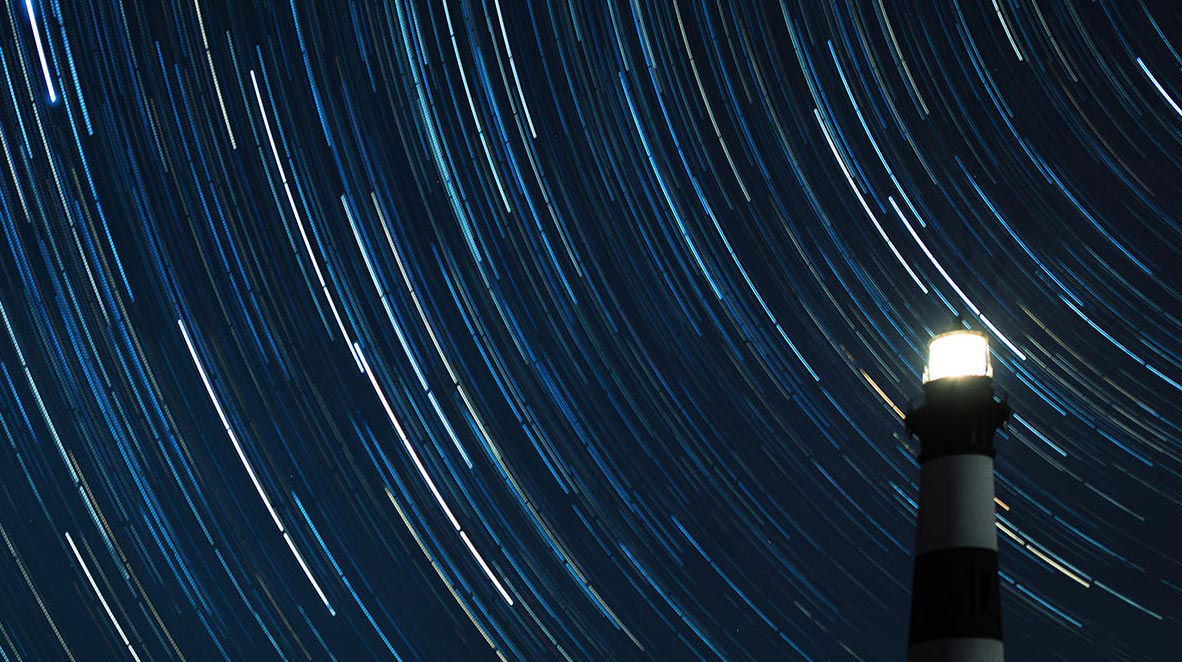 Lighthouse illuminated at night beneath circular star trails in a dark blue sky.
