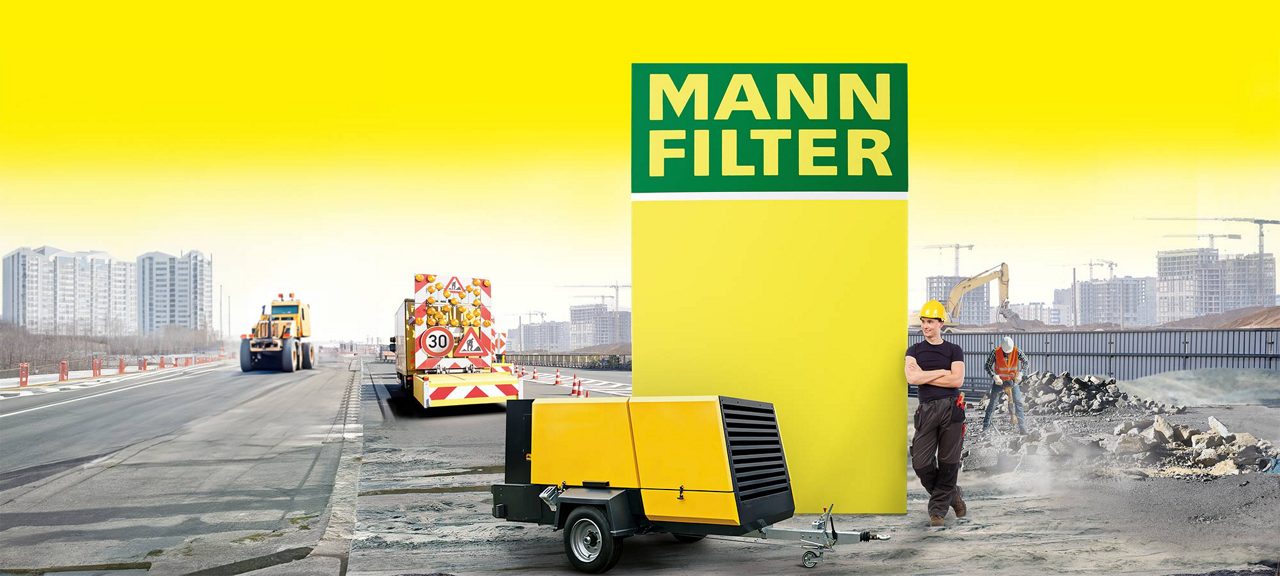 A portable compressor on a worksite with two workers and a bright yellow MANN-FILTER logo