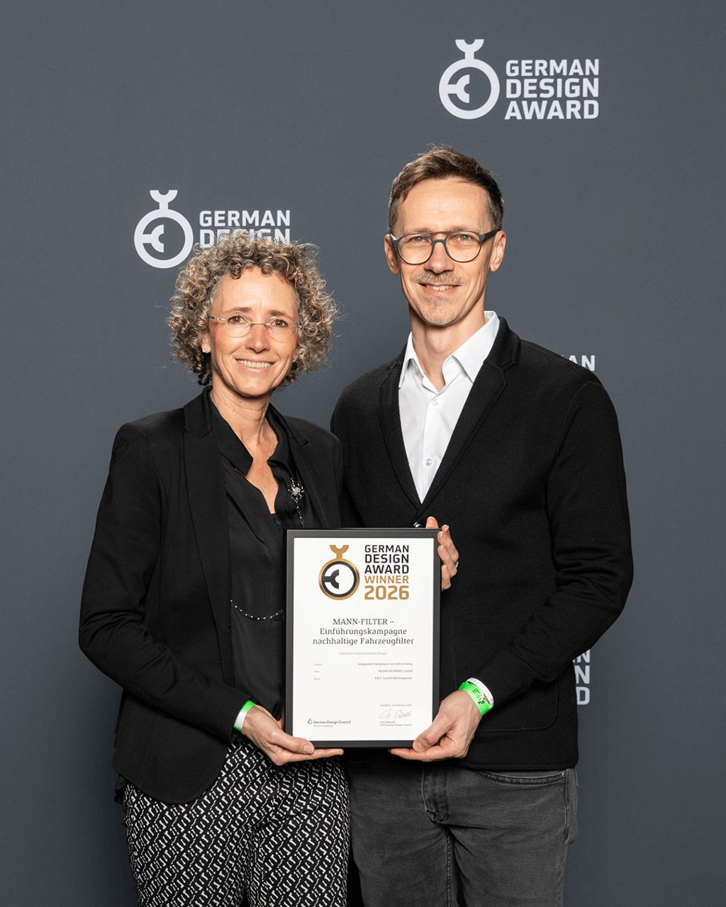 Two people stand in front of a grey backdrop holding the German Design Award 2026