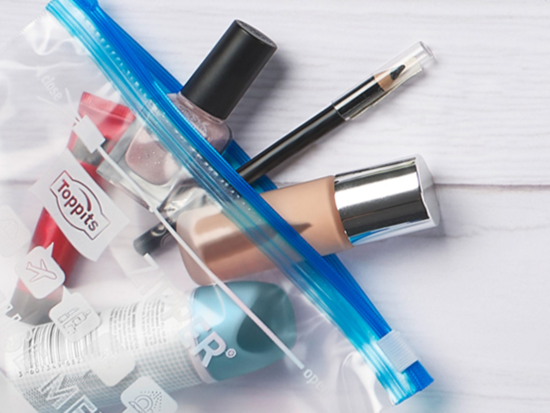 Transparenter Zipper-Beutel von Toppits, gefüllt mit Kosmetikartikeln wie Nagellack, Make-up und einem Pinsel, auf einem weißen Untergrund 