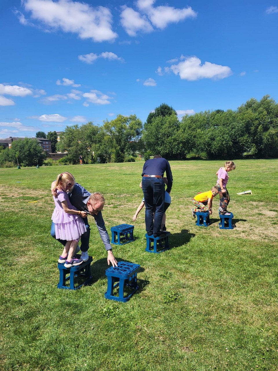 Erwachsene helfen Kindern beim Balancieren über blaue Kunststoffkisten auf einer Wiese, im Hintergrund Bäume und ein weiteres Feld