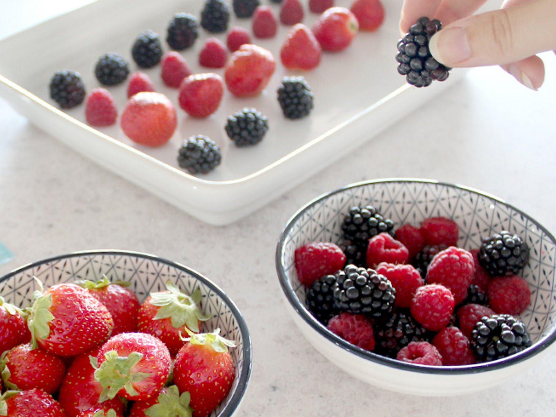 Zwei Schalen mit frischen Erdbeeren, Himbeeren und Brombeeren; im Hintergrund werden Beeren einzeln auf einem weißen Tablett verteilt, eine Hand hält eine Brombeere