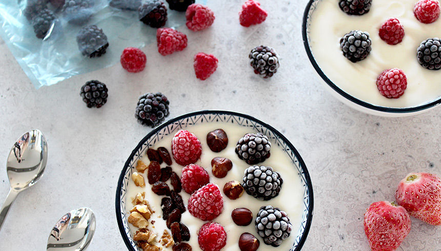 Zwei Schalen mit Joghurt, garniert mit gefrorenen Beeren, Nüssen und Haselnüssen; daneben liegen lose Beeren und zwei Löffel auf einer hellen Oberfläche 