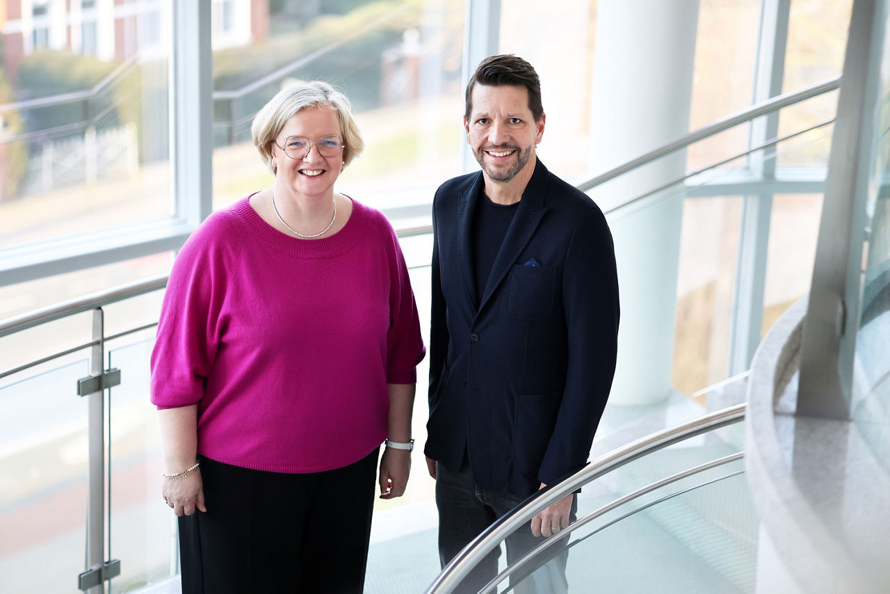 zwei Menschen, eine Frau in magentafarbenem Oberteil und dunkler Hose und ein Mann in dunklem Anzug stehen in einem hell erleuchteten TReppenhaus und blicken zum Betrachter auf. 