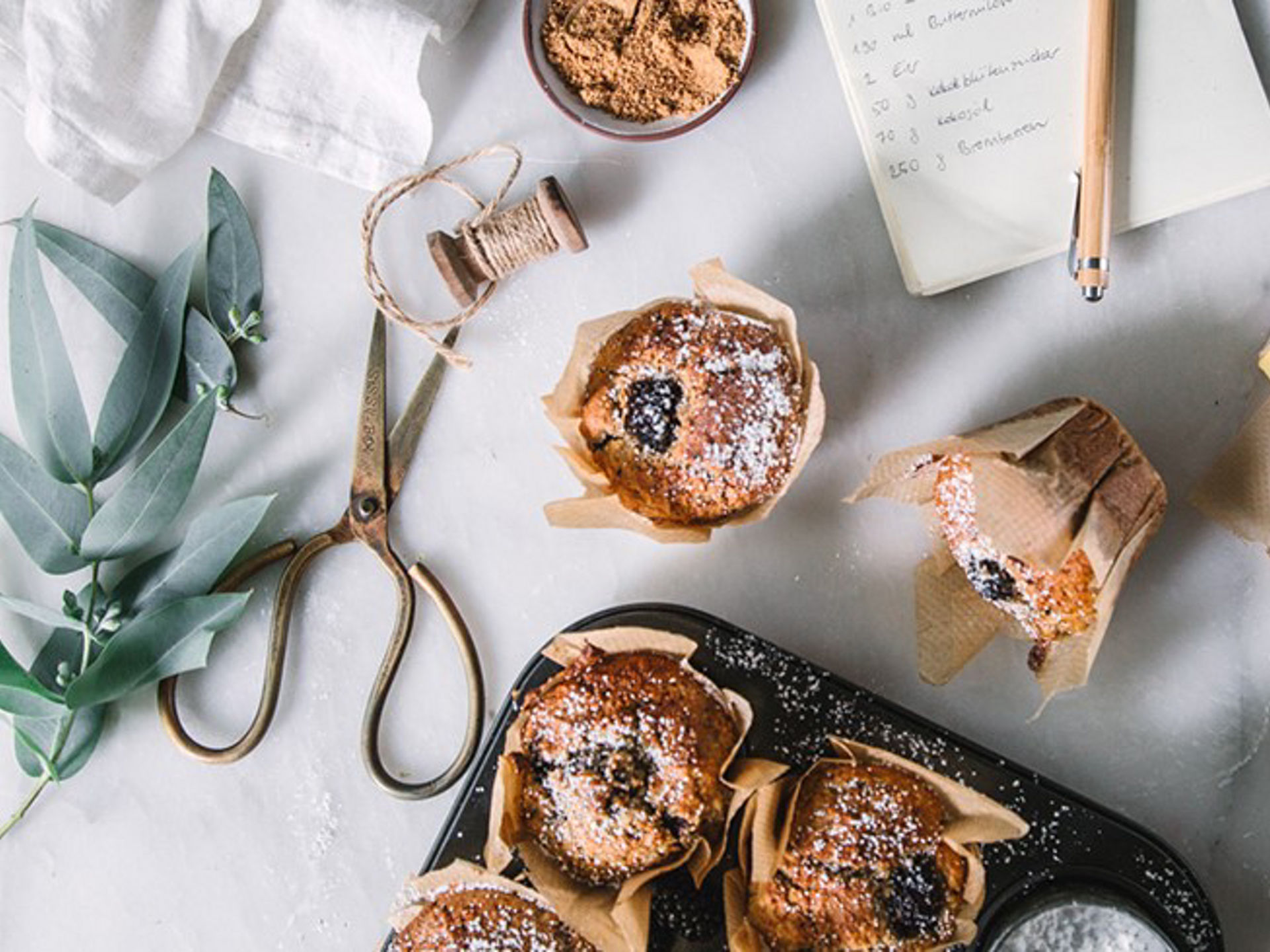 Verschiedene frisch gebackene Muffins in braunen Papierförmchen stehen auf einem Tisch, daneben Backpapier von Toppits, eine Schere, ein Notizbuch mit Rezept und Zutaten wie brauner Zucker