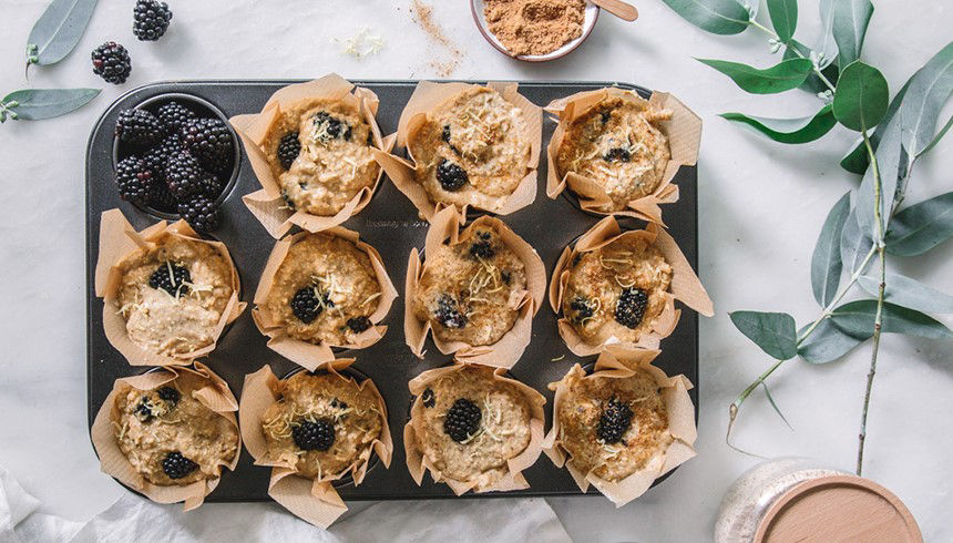 Ein Muffinblech mit zwölf Muffins in braunen Papierförmchen, belegt mit Brombeeren. Daneben eine Schale mit Brombeeren und etwas brauner Zucker. 